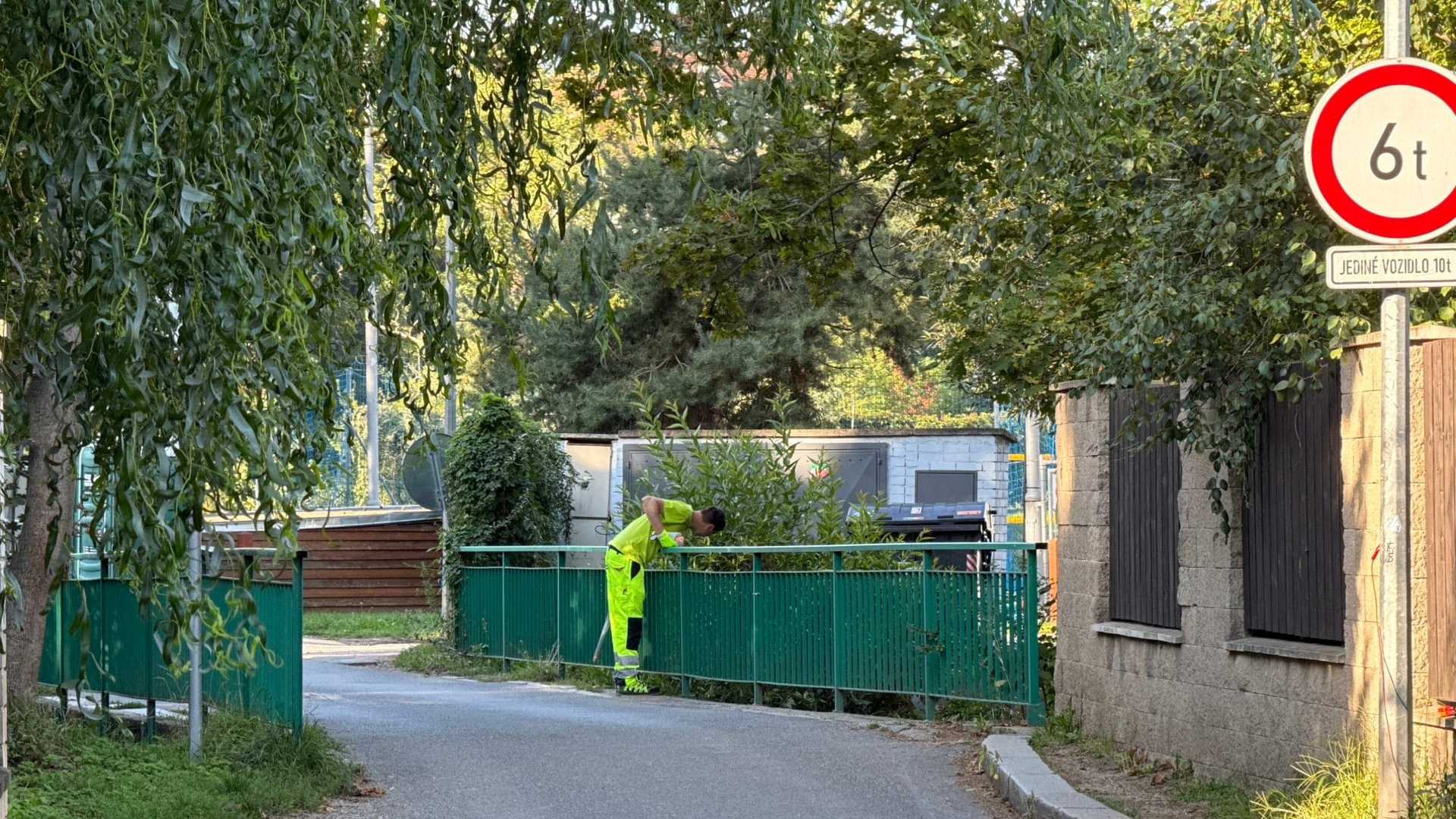 Úzký příjezd k mostu v ulici K Prádelně: jednopruhová asfaltová cesta pod větvemi vrby, podél koryta Botiče zelené ocelové zábradlí, přes které se sklání pracovník v reflexní vestě, na vozovce nastříkané růžové vytyčovací značky a vpravo značka omezující hmotnost na šest tun.
