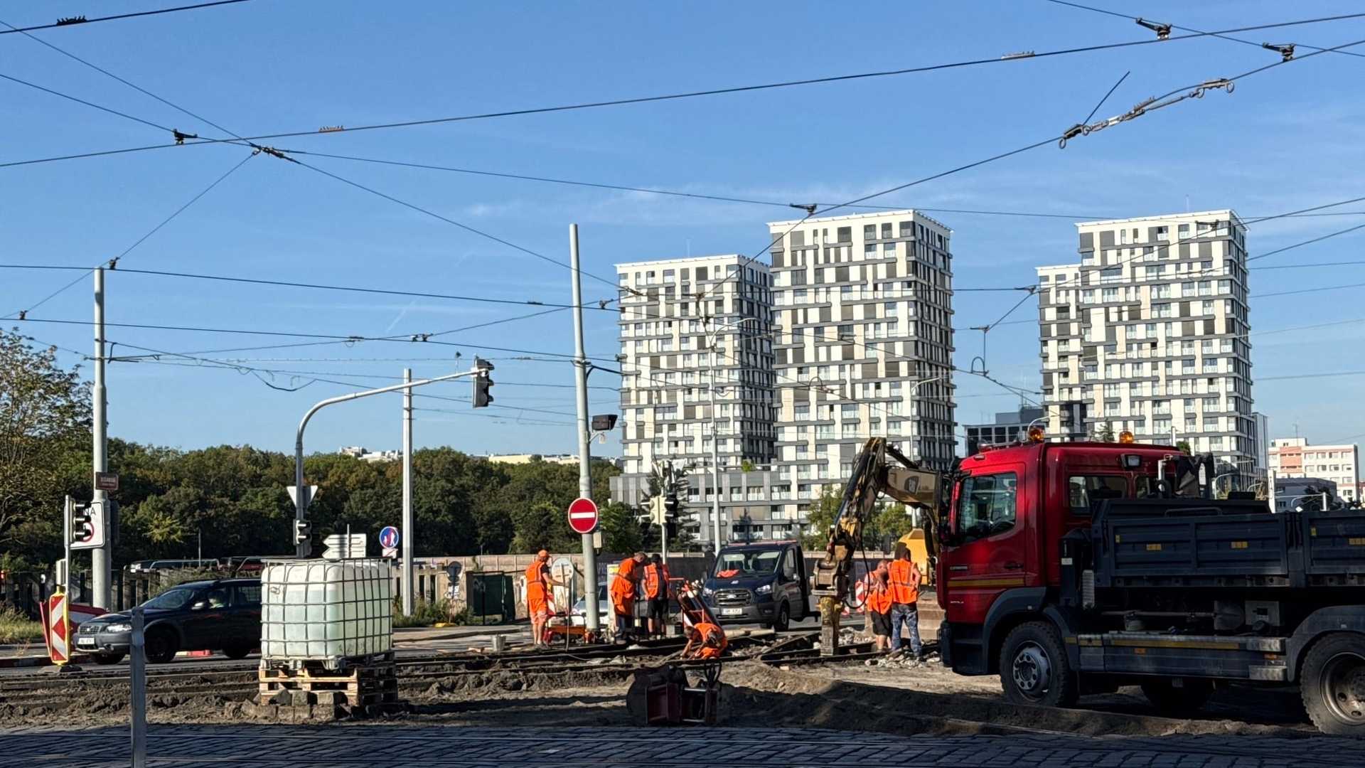 Práce na kolejišti u křižovatky Olšanská: rozebraný tramvajový svršek až na písek a štěrk, parta v oranžových vestách u řezačky kolejnic, rameno žlutého bagru a červený sklápěč, nad tím trolejové vedení a v pozadí tři bílé obytné věže.