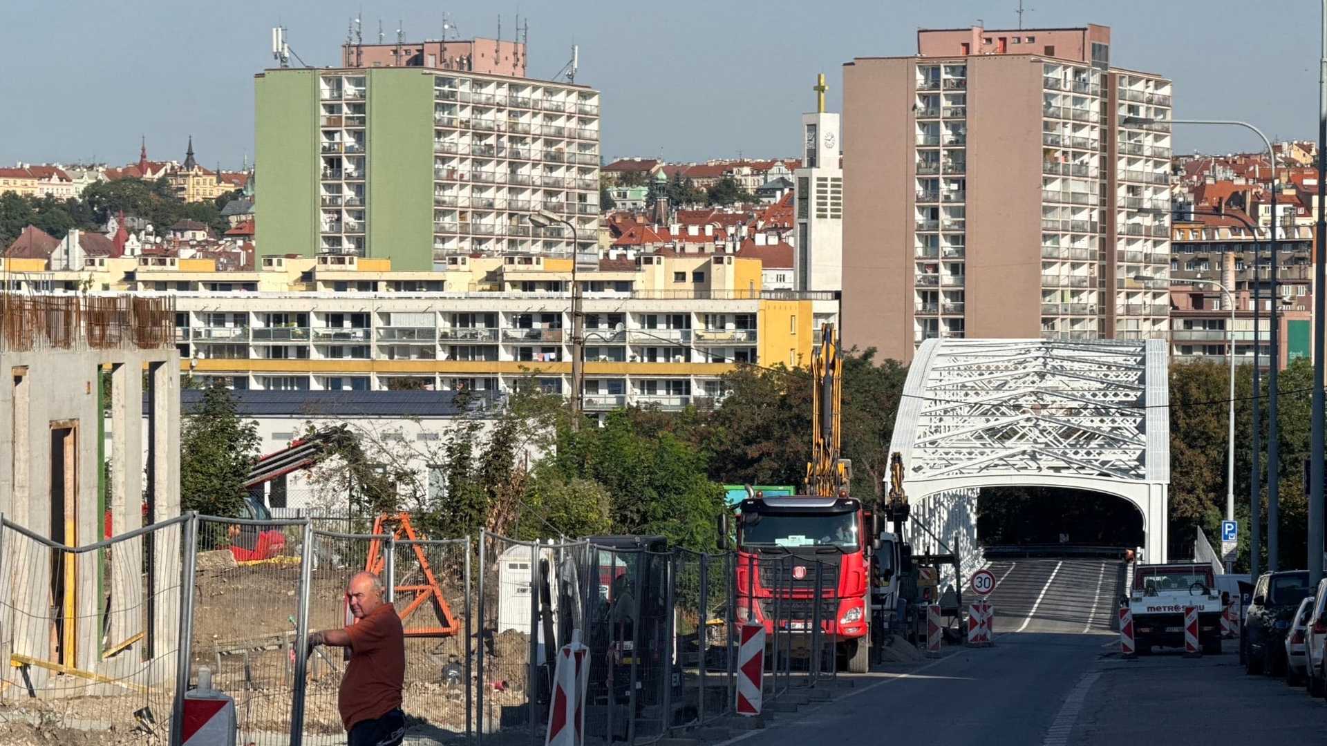 Bíle natřená nýtovaná příhradová konstrukce nadjezdu v Moskevské při pohledu podél vozovky, u vjezdu značka 30 km/h, v popředí oplocená stavba, červený sklápěč a pásový bagr, za tím vršovické výškové domy.