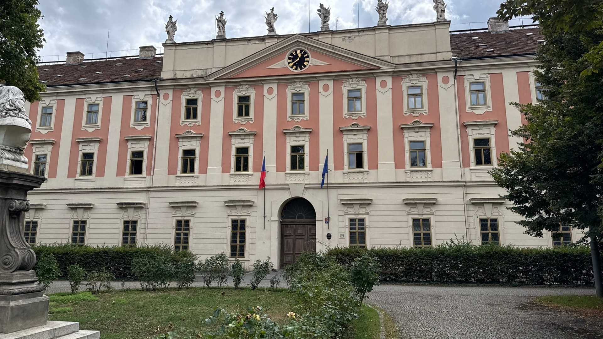 Barokní hlavní průčelí Invalidovny: tři podlaží členěná růžovými a krémovými pilastry, uprostřed tympanon s hodinami se zlatými číslicemi, na atice šest kamenných soch a u klenutého vstupu česká a unijní vlajka.