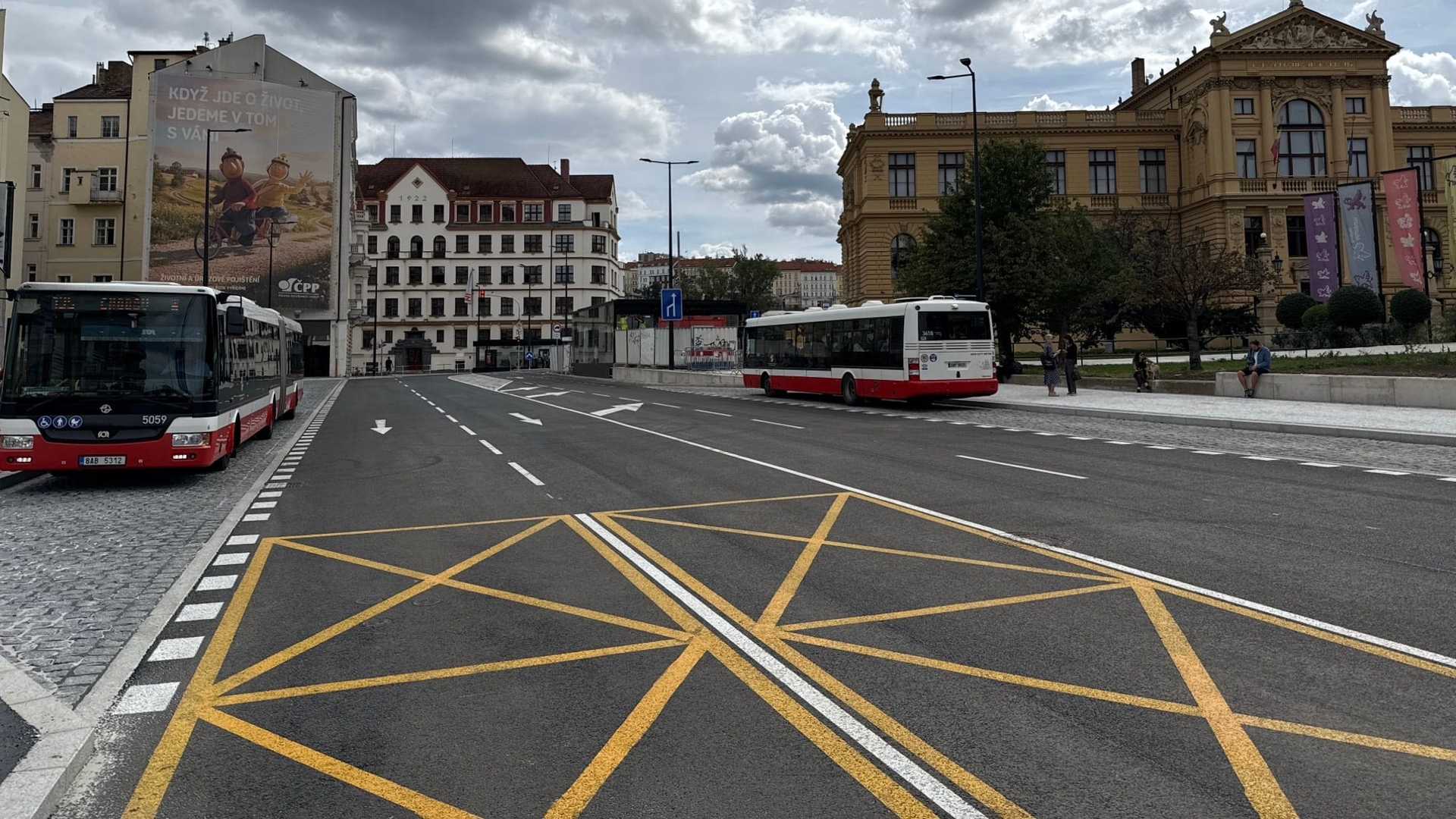 Znovu otevřená ulice Ke Štvanici s novým asfaltem, bílým vodorovným značením a žlutou křižovatkovou plochou, u levého obrubníku stojí kloubový autobus DPP a druhý odjíždí, v pozadí žlutá novorenesanční budova a stavební oplocení.