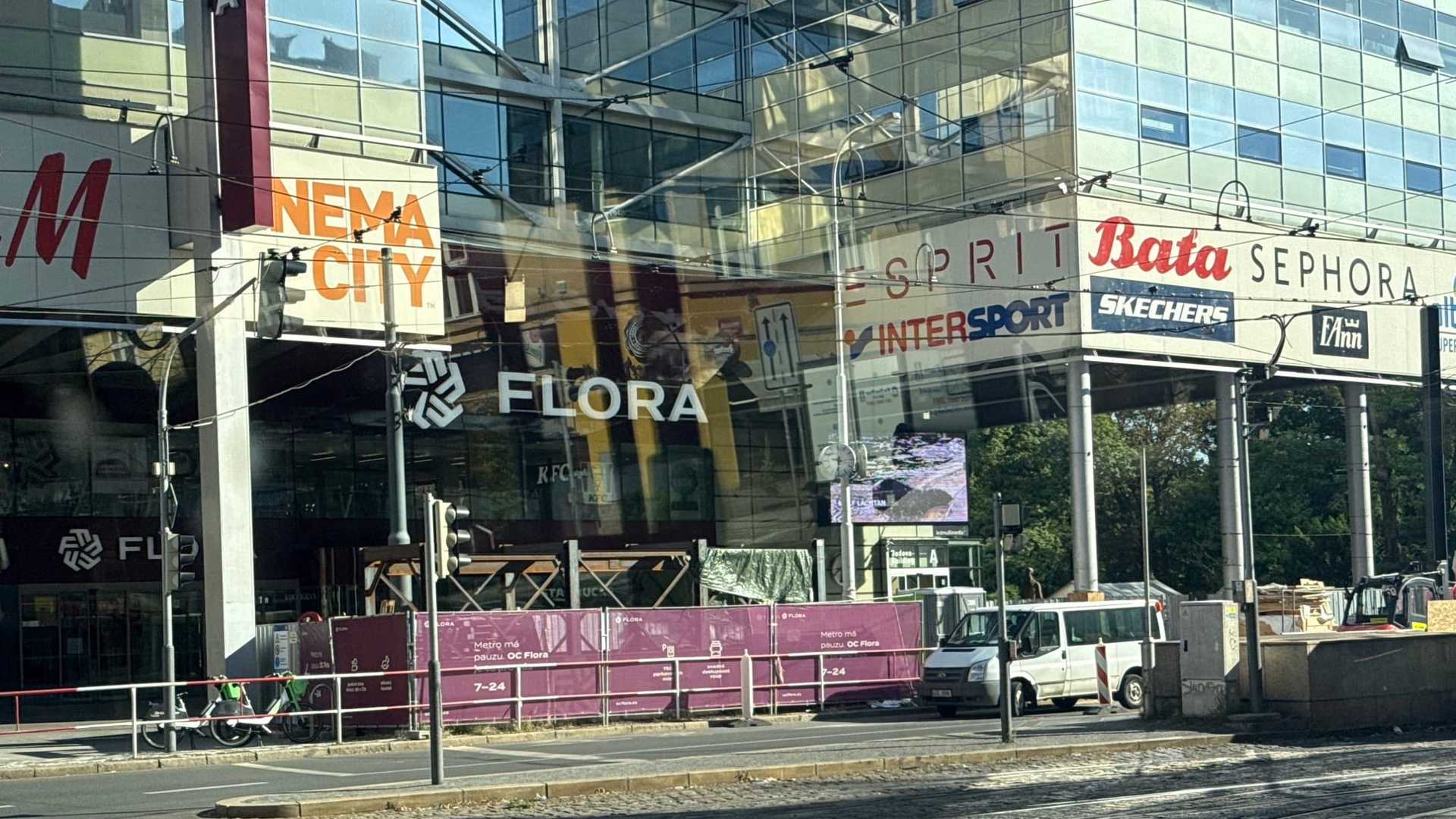 Obchodní centrum Flora nad uzavřeným vstupem do metra, přes vestibul purpurové ohrazení s nápisem Metro má pauzu, vedle zaparkovaná bílá dodávka, za ním technika a v popředí zatáčející tramvajové koleje.
