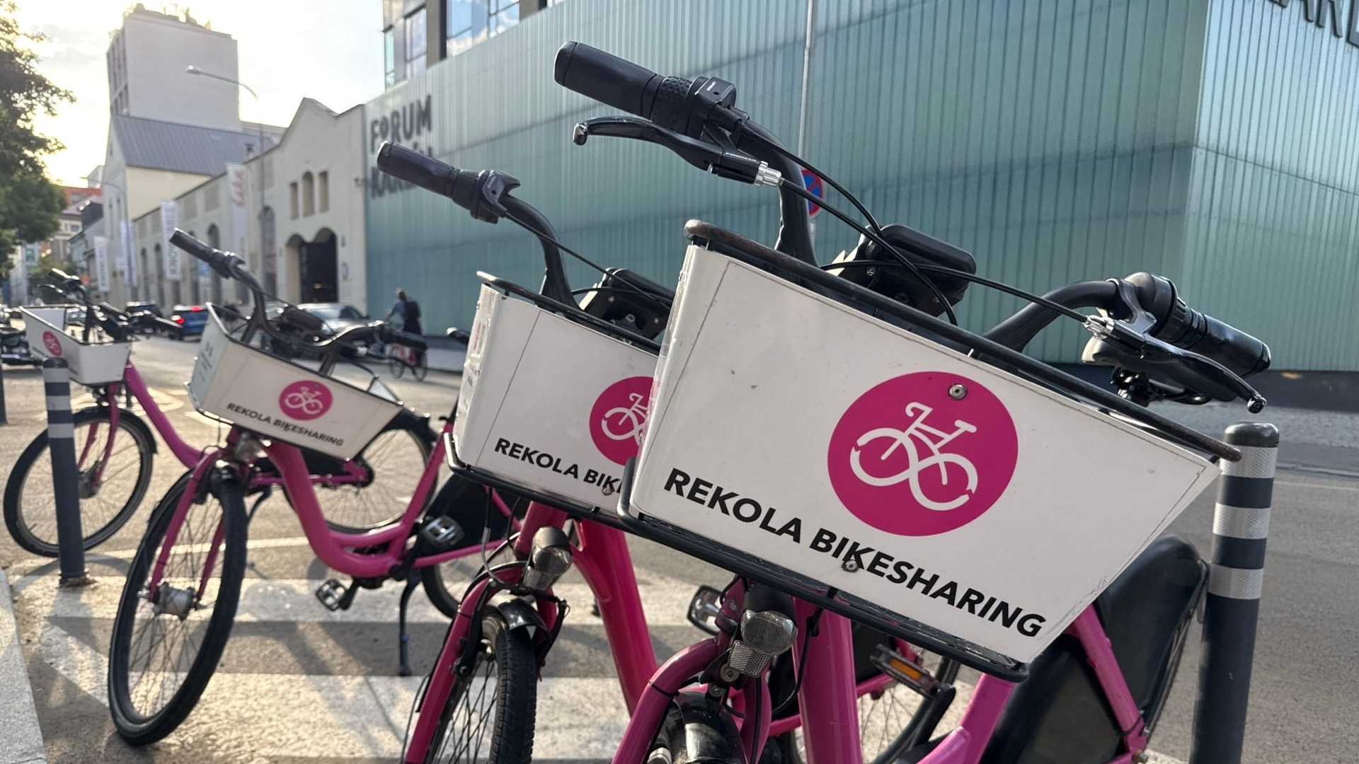 Tři růžová kola Rekola s bílými přepravkami a purpurovým logem provozovatele, zaparkovaná na přechodu u sloupku, za nimi nápis Forum Karlín a světle zelená vlnitá fasáda.