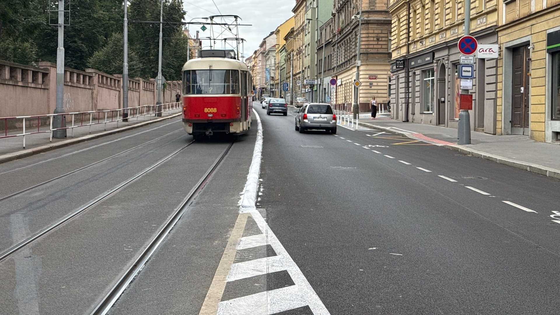Krémovočervená tramvaj T3 s číslem 8088 na vyhrazeném pásu v Plzeňské, vedle ní čerstvé bílé značení a symbol kola na novém povrchu vozovky, před ní auta a vpravo řada nájemních domů.