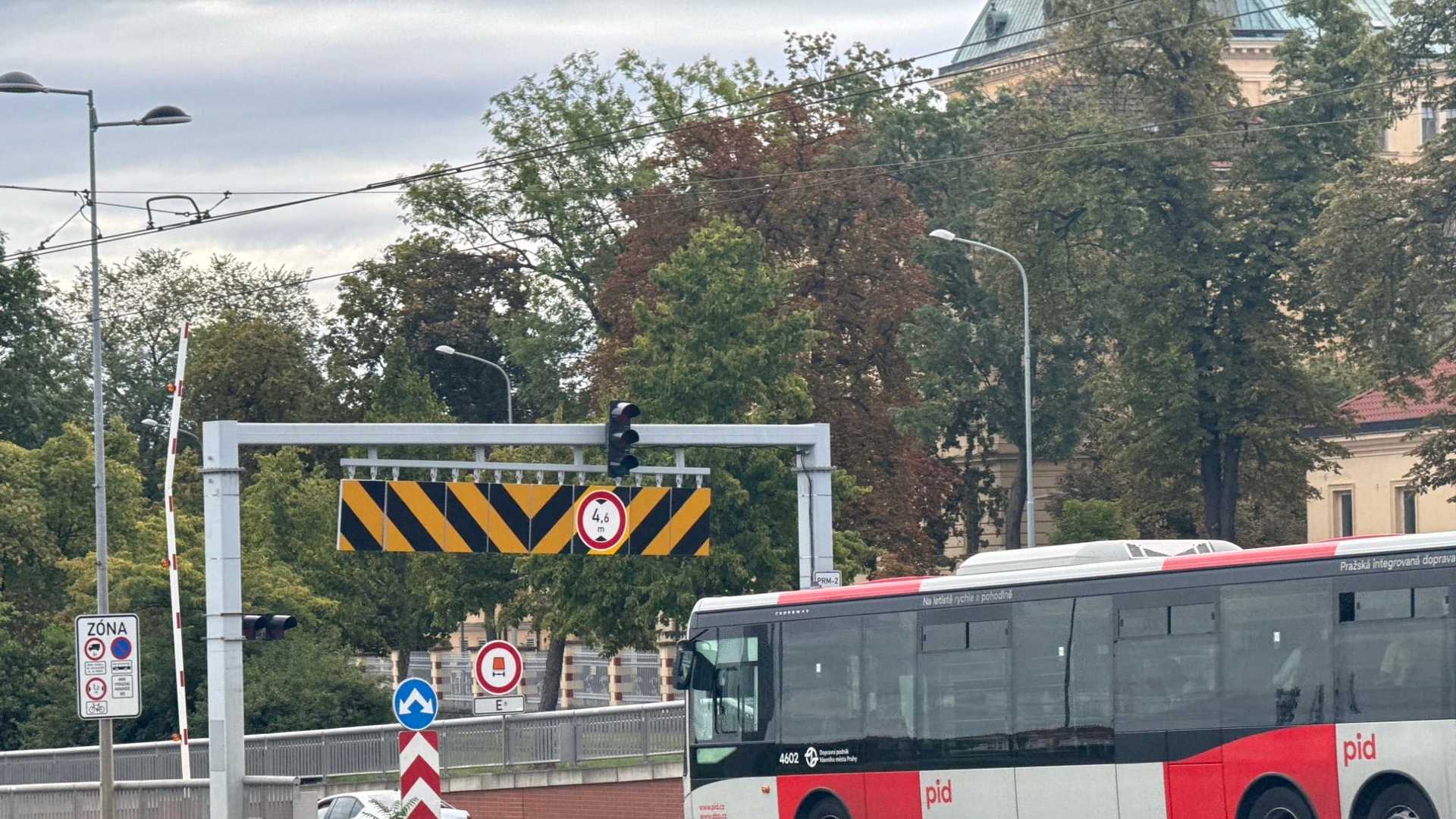 Červenobílý autobus PID projíždí kolem tunelového portálu: nad vozovkou ocelový portál s černožlutými šipkami a značkou omezující výšku na 4,6 metru, vedle zvednutá závora, za tím stromy a světlá historická budova.