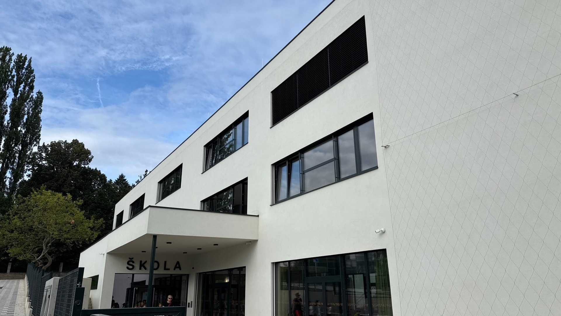 Nový bílý školní pavilon při pohledu od nároží: tři podlaží s tmavými rámy oken v hladké omítané fasádě, pod vstupní stříškou tmavý nápis ŠKOLA, prosklené přízemí, za nímž jsou vidět děti a dospělí, a na slepém štítu lanový rošt pro popínavou zeleň.