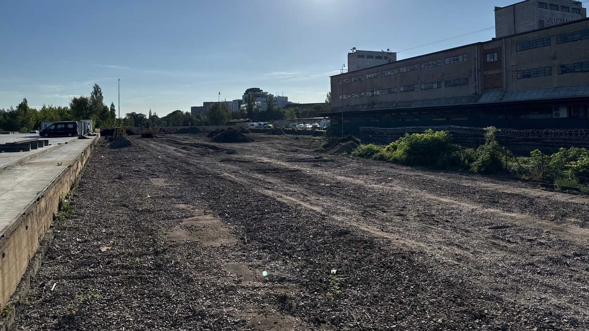 Široký pruh vyklizené a srovnané plochy mířící do dálky, podél něj hromady výkopku, vlevo betonová nákladová rampa a vpravo dlouhá funkcionalistická budova nákladového nádraží s vyloženou stříškou.