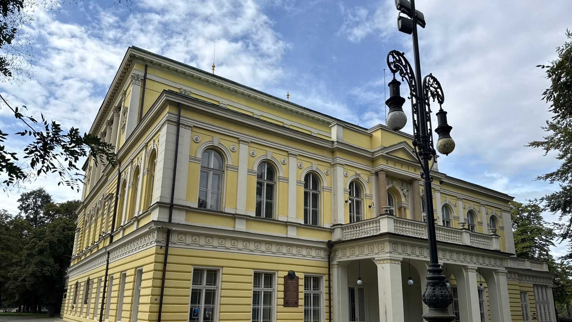 Palác Žofín na Slovanském ostrově od nároží: dvoupodlažní žluto-bílá novorenesanční budova s půlkruhově zakončenými okny, na tympanonu nápis ŽOFÍN nad sloupovým portikem, před ním zdobený litinový kandelábr a po stranách stromy.