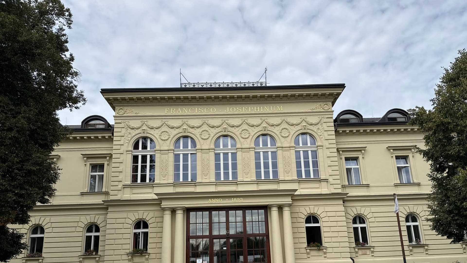 Hlavní průčelí domova Palata: světle žlutá dvoupodlažní budova s nápisem FRANCISCO-JOSEPHINUM nad girlandovou štukovou výzdobou a pěti klenutými okny, nad vstupem ANNO-1893, na předním dvoře fontánka a lavičky, na jedné z nich sedí žena.