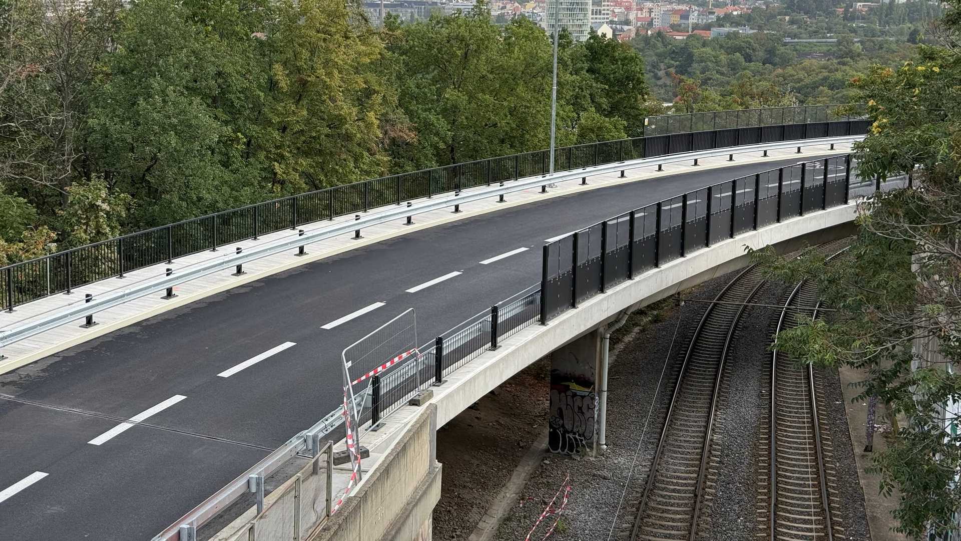 Most Pod Krejcárkem shora od jižního konce: zaoblená betonová mostovka s čerstvým asfaltem a novým vodorovným značením, po obou stranách pozinkované svodidlo a tmavé clony, pod mostem v zářezu železniční trať a za ní až k obzoru severní části Prahy.
